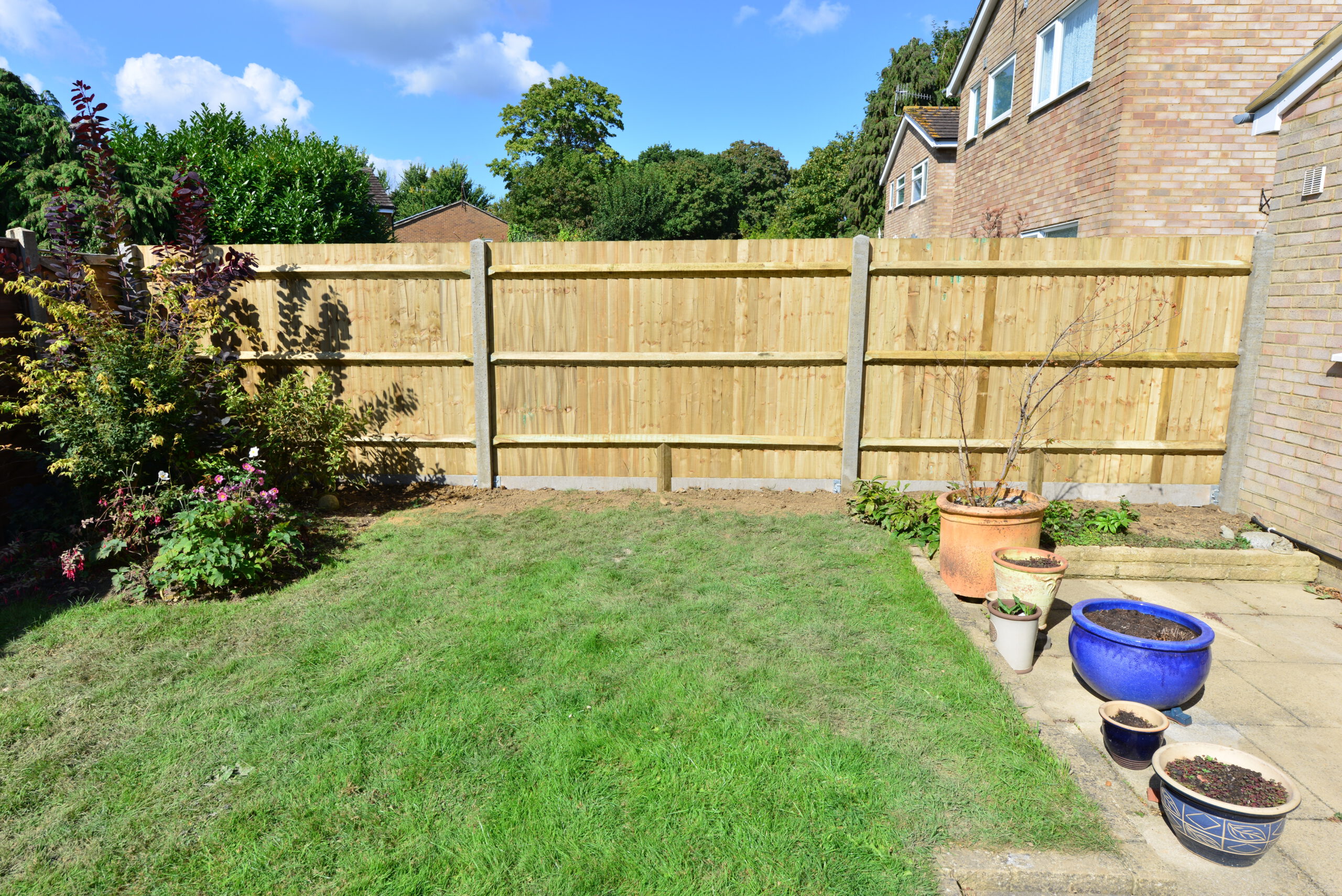 A backyard with new garden fencing, green grass, some shrubs along the left side, and several potted plants on a patio with new paving near a brick house under a blue sky with clouds.