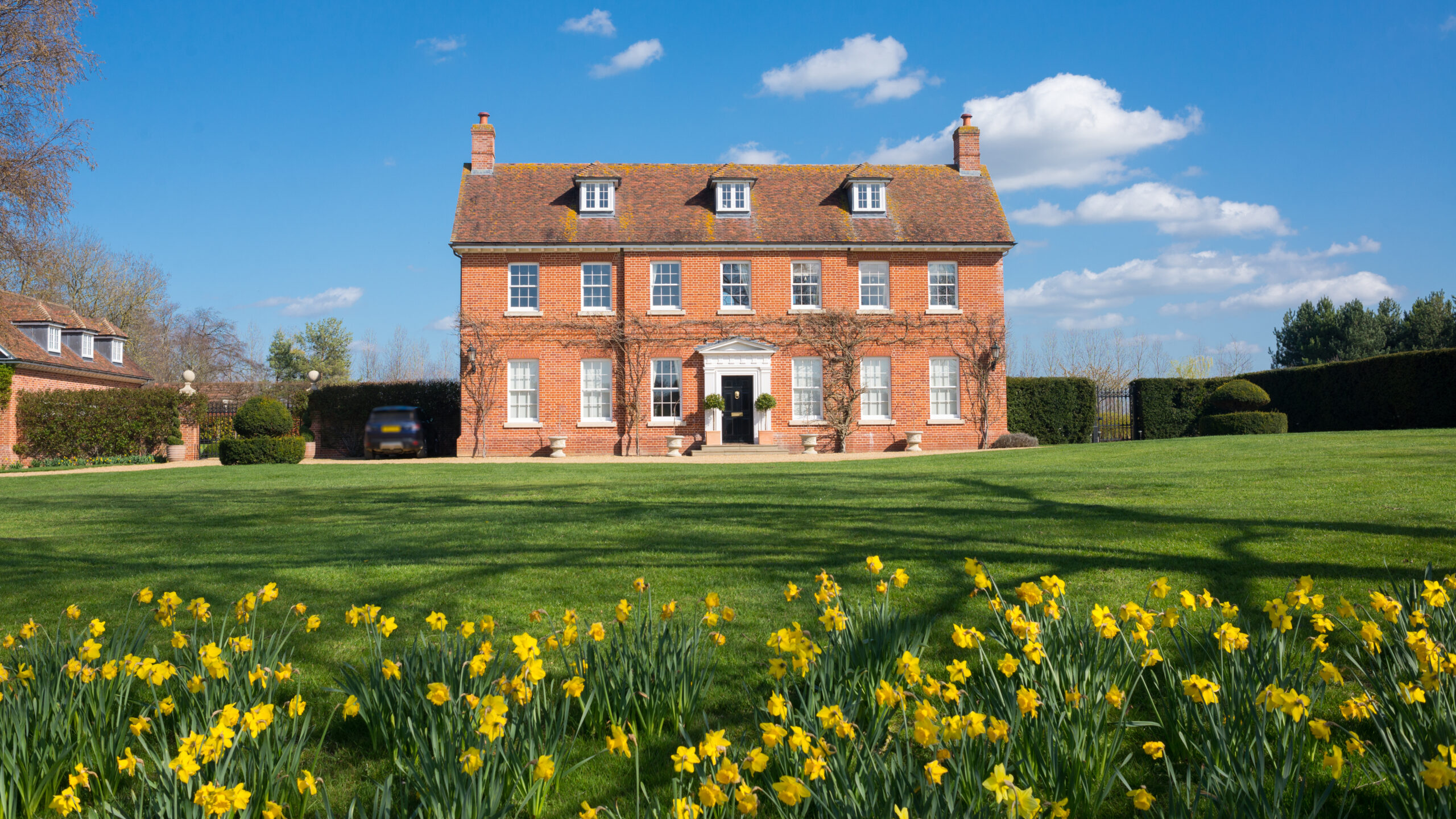 A large red-brick Georgian house with white-framed windows and chimneys stands behind a neatly mowed lawn, where blooming yellow daffodils border elegant timber decking, all beneath a bright blue sky with scattered clouds.