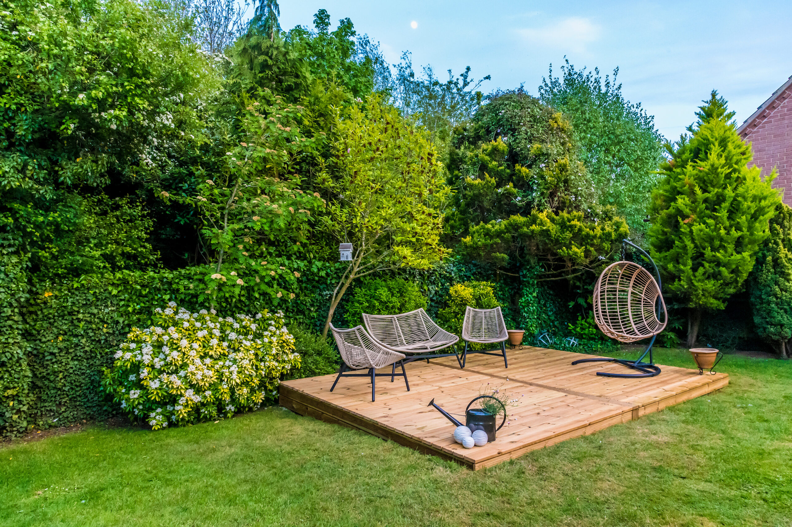 A timber decking area with modern outdoor chairs and a hanging chair sits in a lush, green garden surrounded by dense bushes and trees. A watering can and garden tools rest on the edge, showcasing expert landscaper touches.