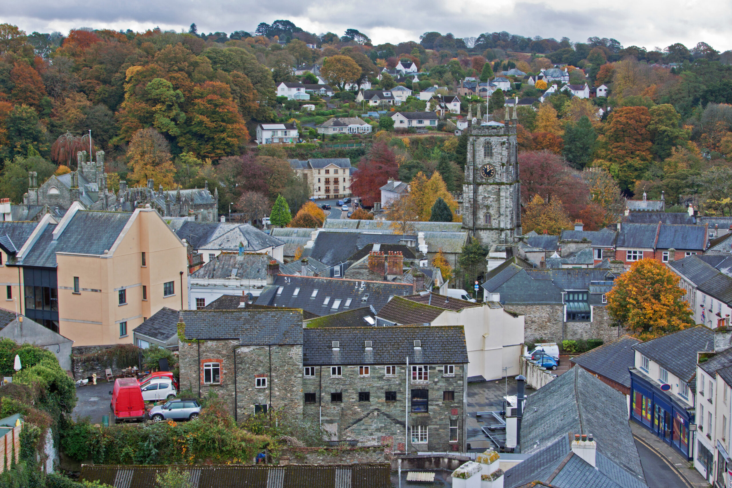 A scenic view of a small town with stone buildings, a church tower with a clock, and colorful autumn trees on the hills in the background. Neatly trimmed garden fencing hints at the work of a skilled landscaper. The sky is overcast.