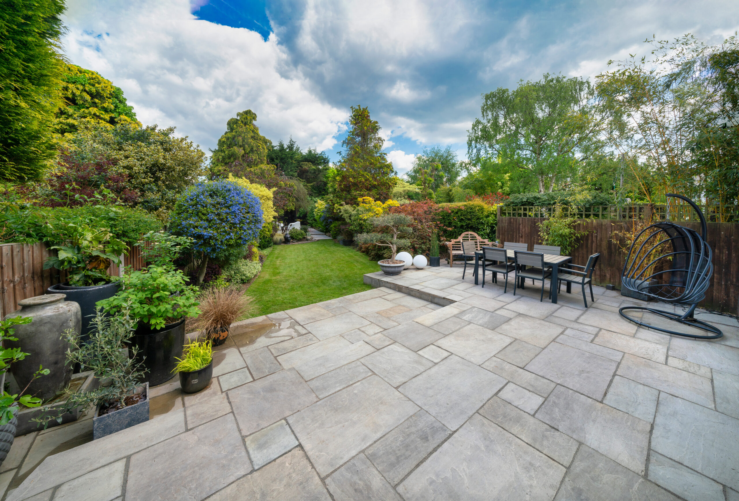 Spacious backyard with a large stone patio, outdoor dining table and chairs, potted plants, and a green lawn bordered by colorful shrubs, trees, and timber decking under a partly cloudy sky.