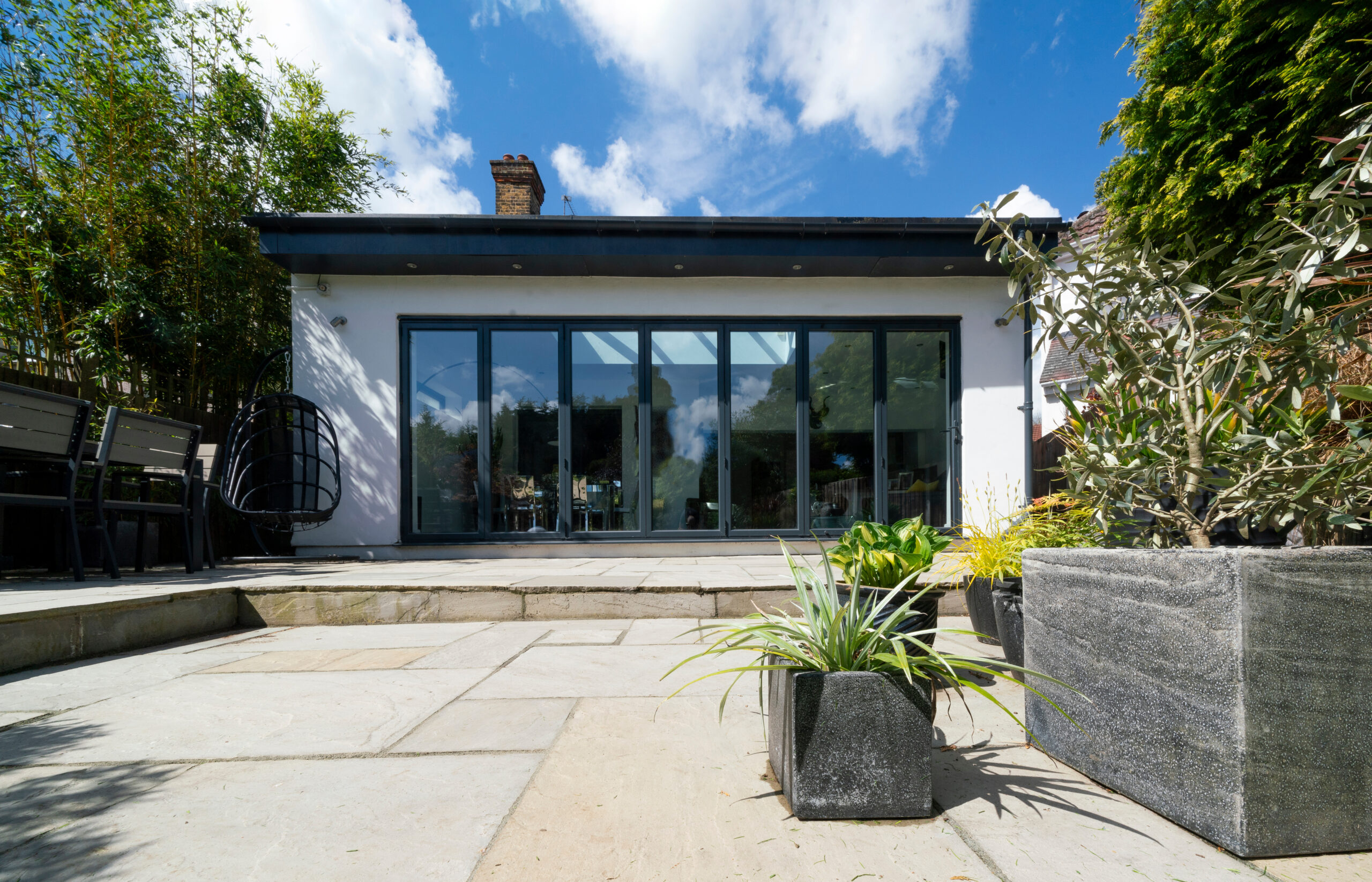 A modern white house with large glass doors opening onto a stone patio with new paving, outdoor dining area, and several potted plants, set against a blue sky with scattered clouds.